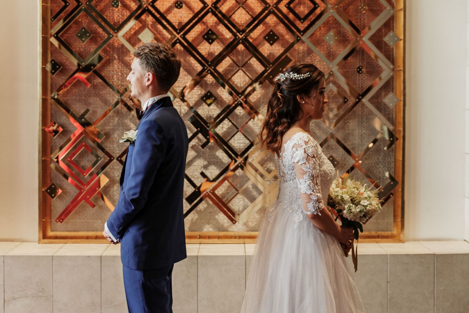 Novios posan espalda con espalda ante panel decorativo dorado; él traje azul, ella vestido encaje con ramo.