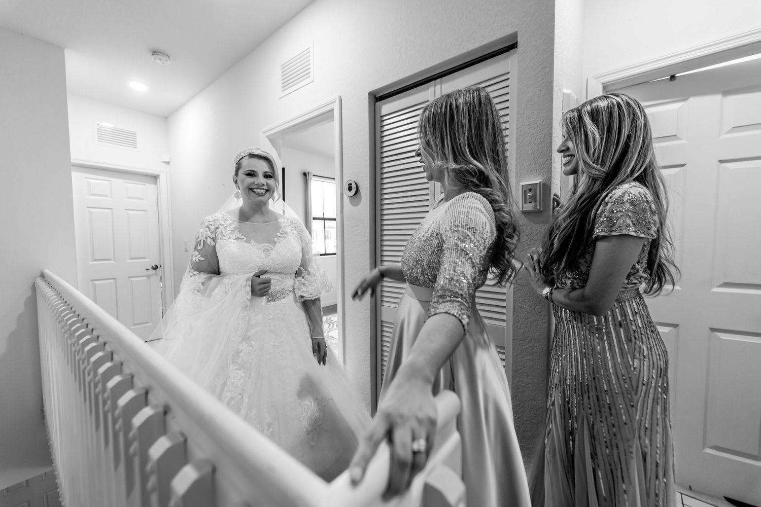 Foto B&W: Novia sonriente muestra su vestido a damas de honor admiradas en pasillo interior. Reacción boda.