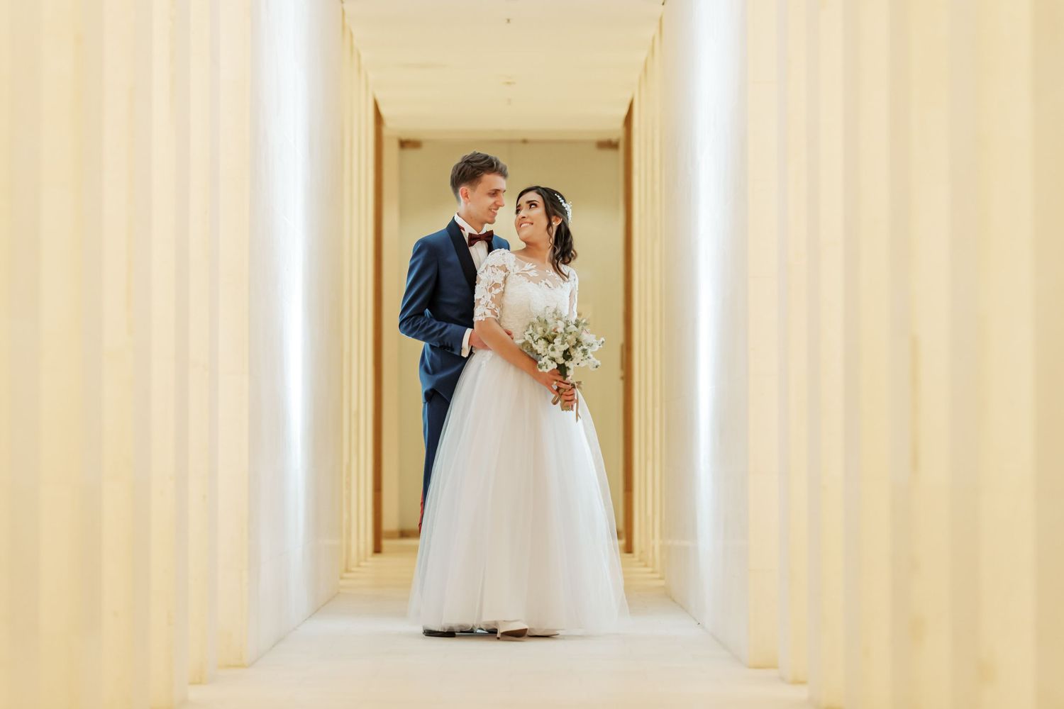 Novios se miran posando en pasillo estrecho y luminoso; ella vestido blanco manga larga, él traje azul. Boda.