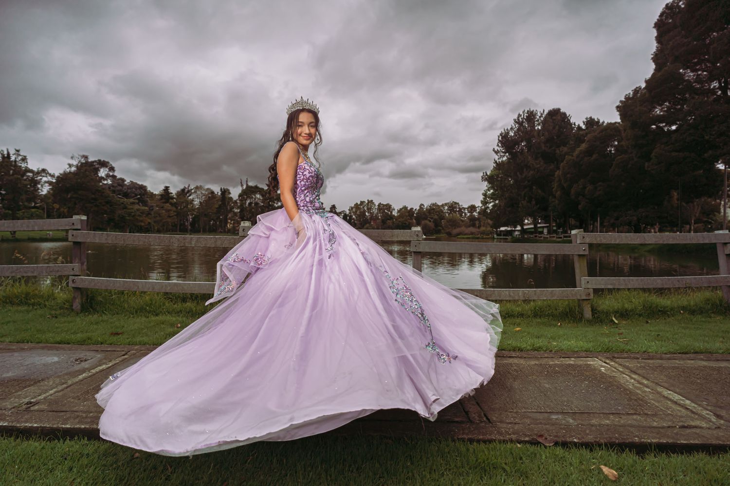 Quinceañera con vestido lavanda gira feliz junto a lago bajo cielo nublado; sesión fotos exterior.