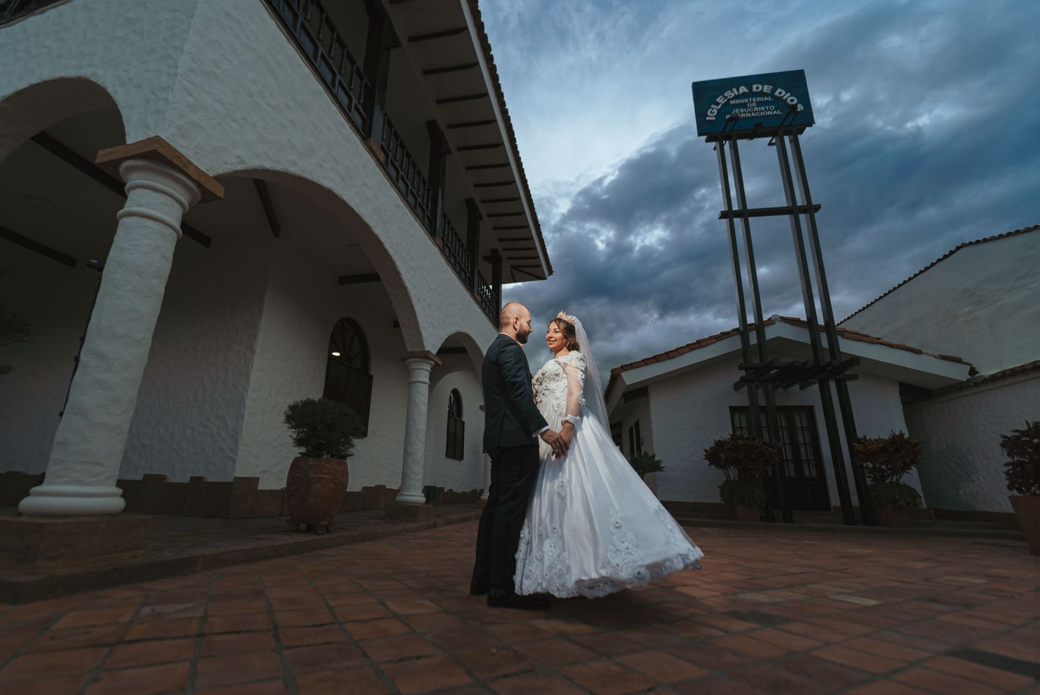 Novios se miran románticamente tomados de la mano frente a edificio 'Iglesia de Dios'; cielo nublado. Boda IDMJI.