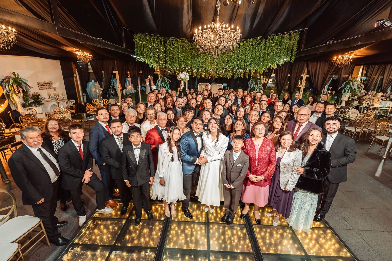 Foto grupal boda: Novios y numeroso grupo de invitados posan elegantes en salón recepción con pista iluminada.