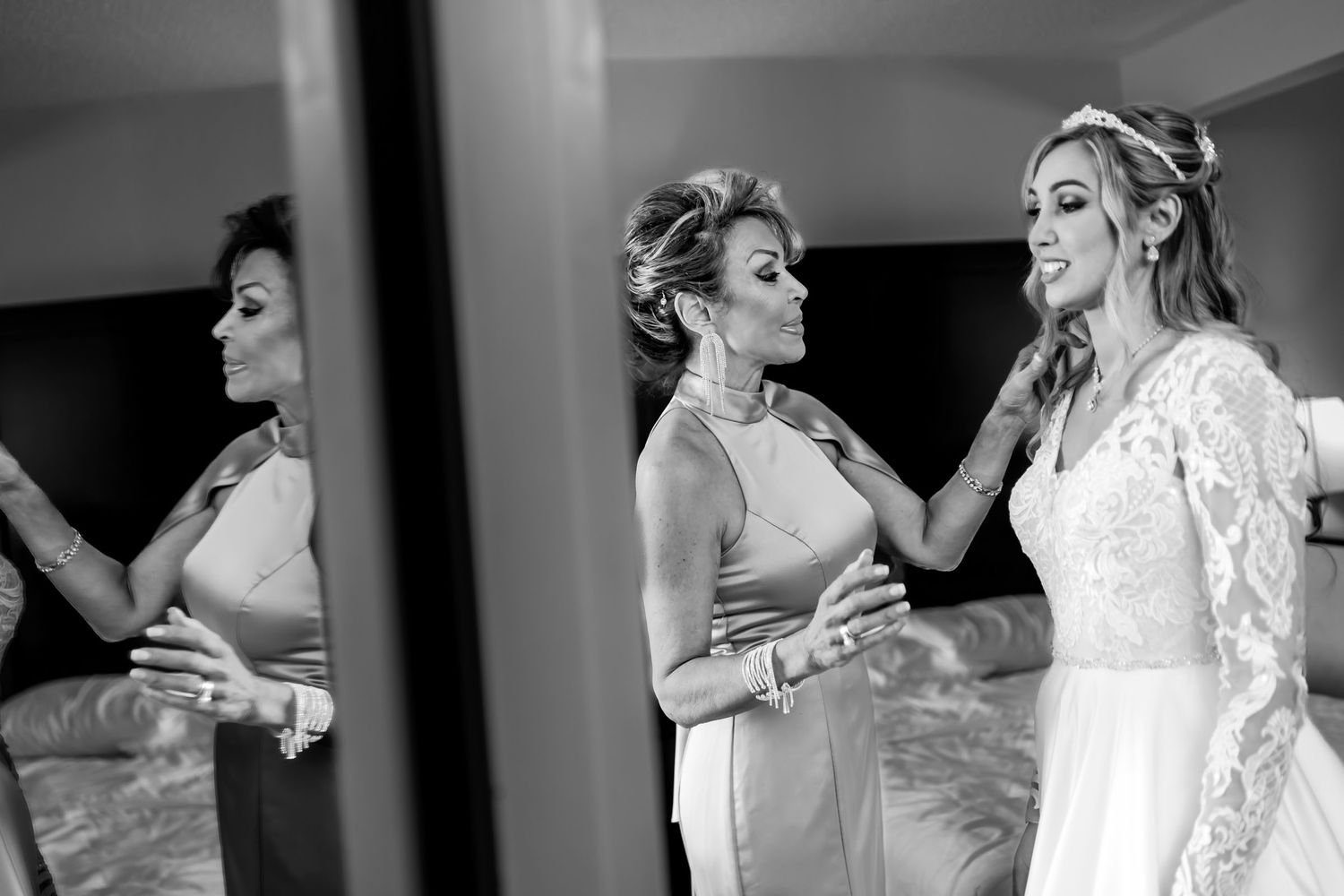 Foto B&W: Madre ayuda a novia (vestido encaje) a prepararse frente a espejo; momento emotivo boda.