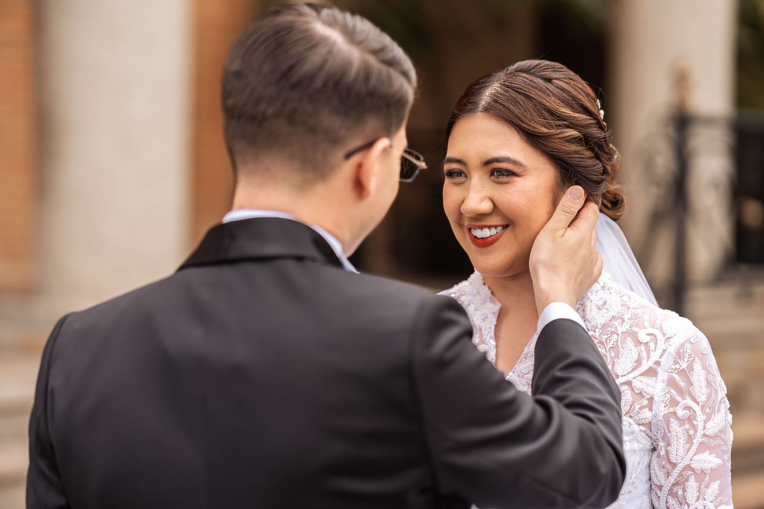 Novio acaricia tiernamente mejilla de novia sonriente; él con esmoquin, ella con vestido encaje y velo. Foto de boda.
