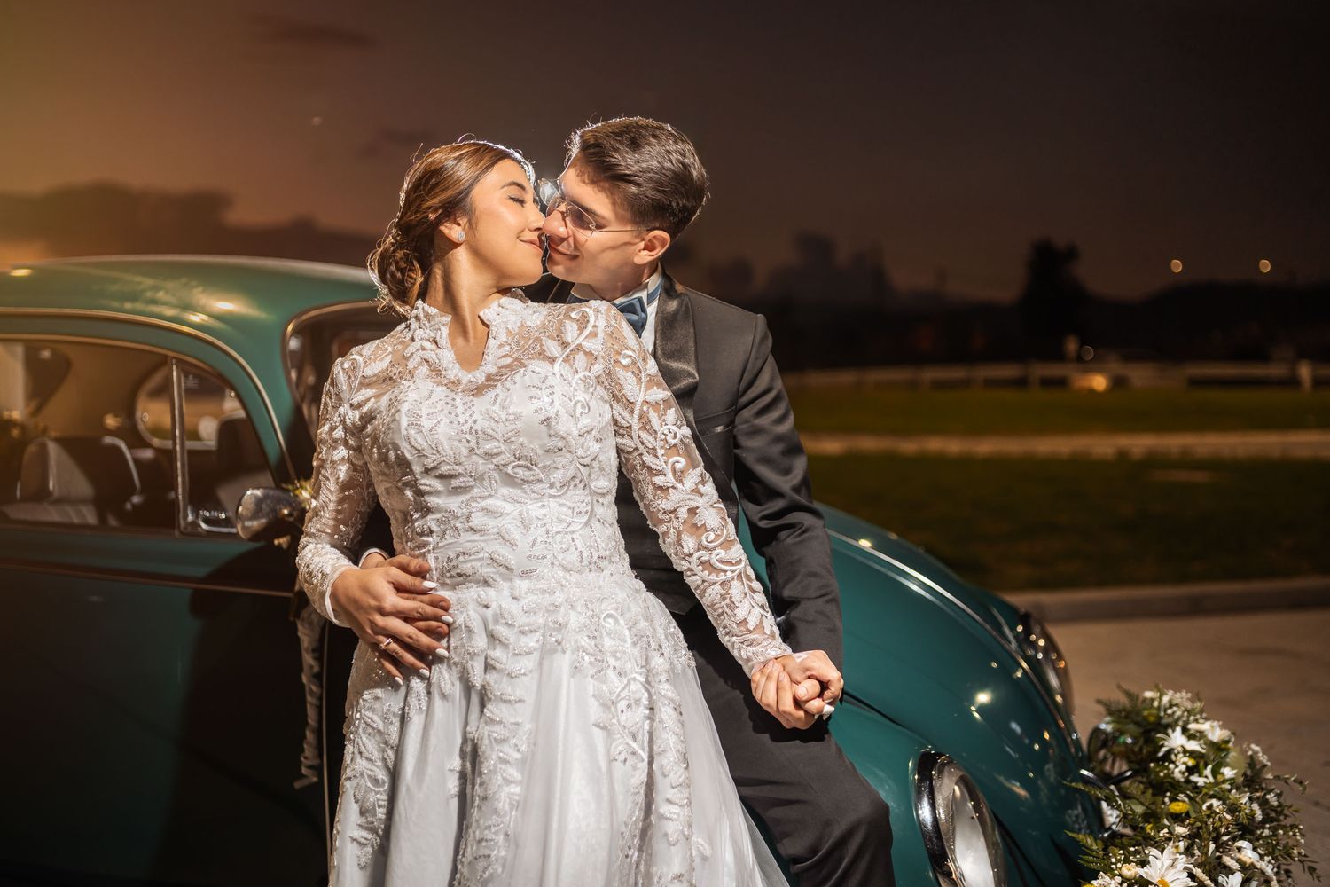Novios se besan apasionadamente junto a coche verde clásico en la noche. Foto de boda exterior romántica.