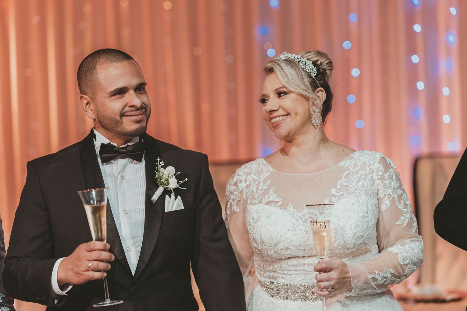 Novios sonrientes se miran sosteniendo copas de champagne durante brindis en recepción de boda; fondo cortina luces.