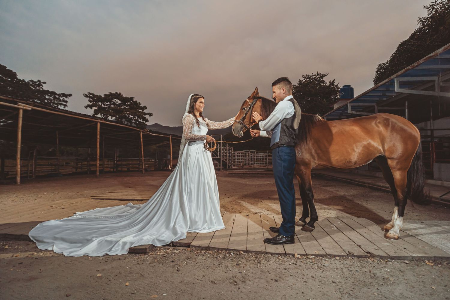 Pareja de novios interactúa con un caballo en un entorno rural. Foto de boda campestre con cielo de fondo al atardecer