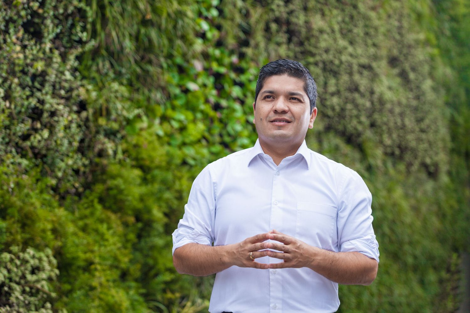 Hombre profesional con camisa blanca, manos juntas, posa sonriente frente a jardín vertical. Retrato exterior.