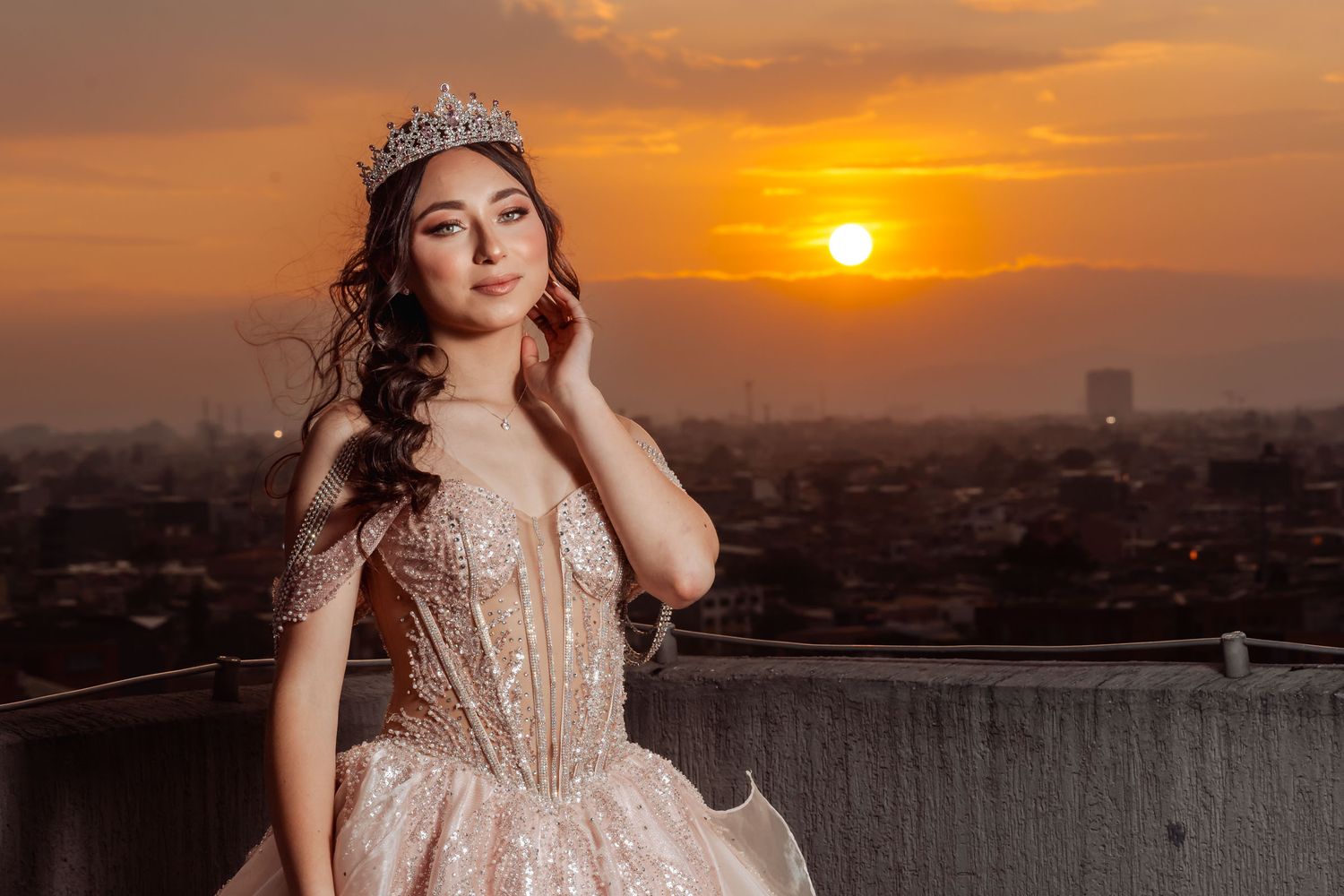Retrato quinceañera con vestido rosa corsé y tiara en terraza; sol poniéndose sobre ciudad brumosa, luz dorada.