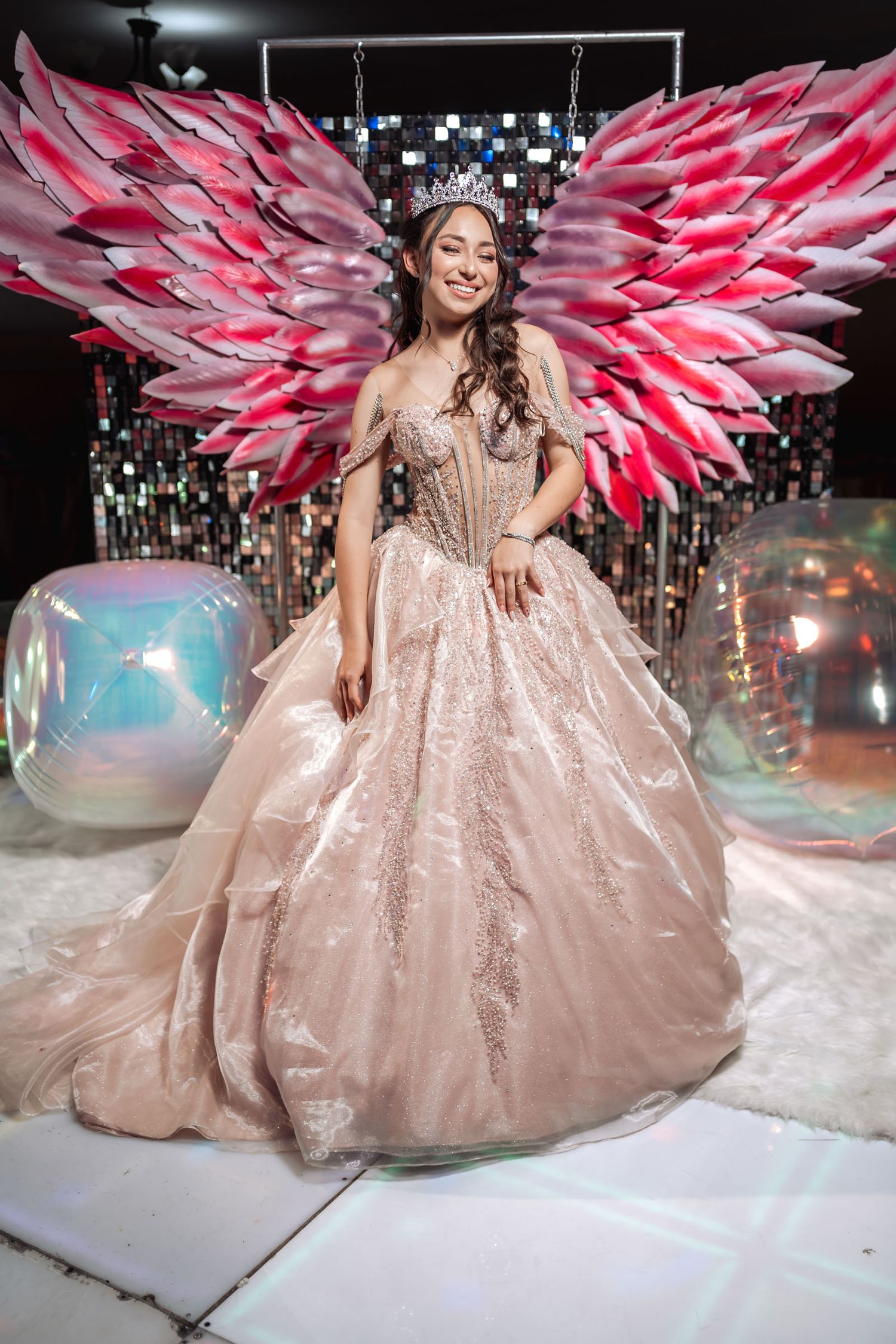 Quinceañera sonriente con vestido princesa rosa y tiara posa con grandes alas de ángel iluminadas; fiesta 15.