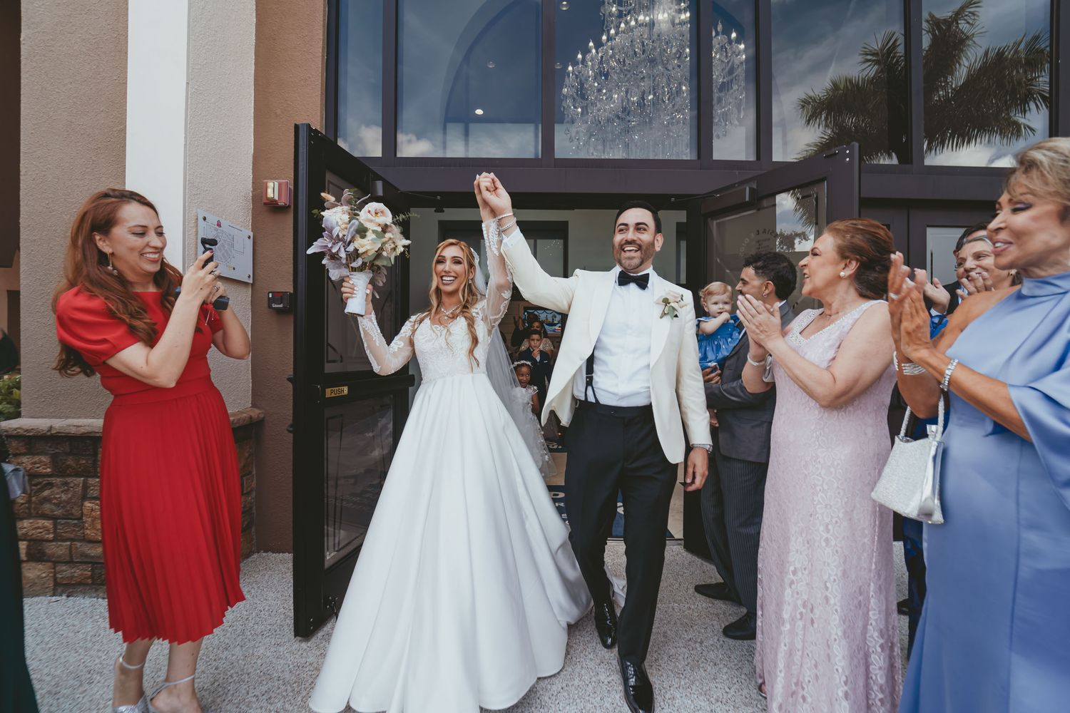 Novios felices salen de iglesia con brazos alzados; invitados aplauden. Salida nupcial boda.