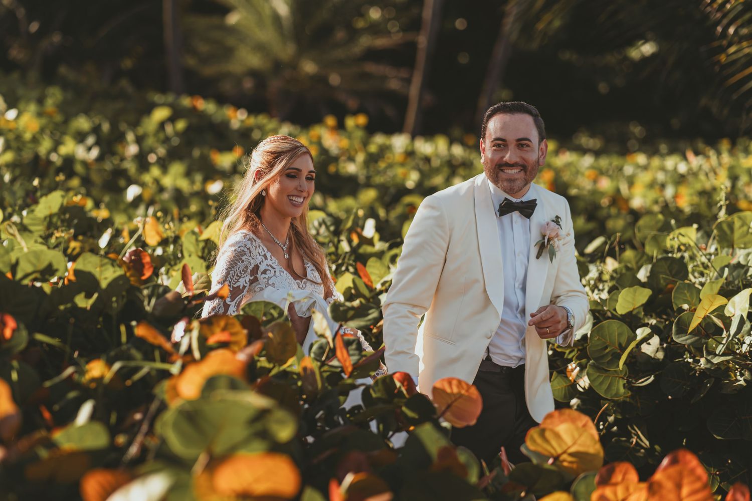Novios sonrientes caminan entre arbustos con flores naranjas; él chaqueta clara. Boda destino tropical.