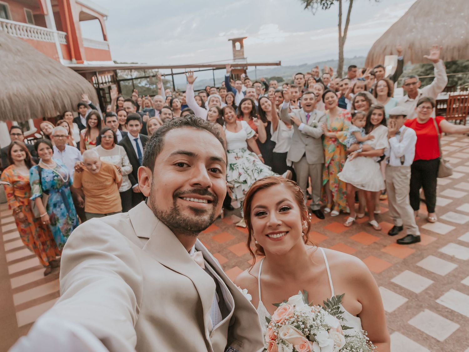 Novios toman selfie sonrientes con gran grupo de invitados en terraza exterior; ambiente festivo boda.