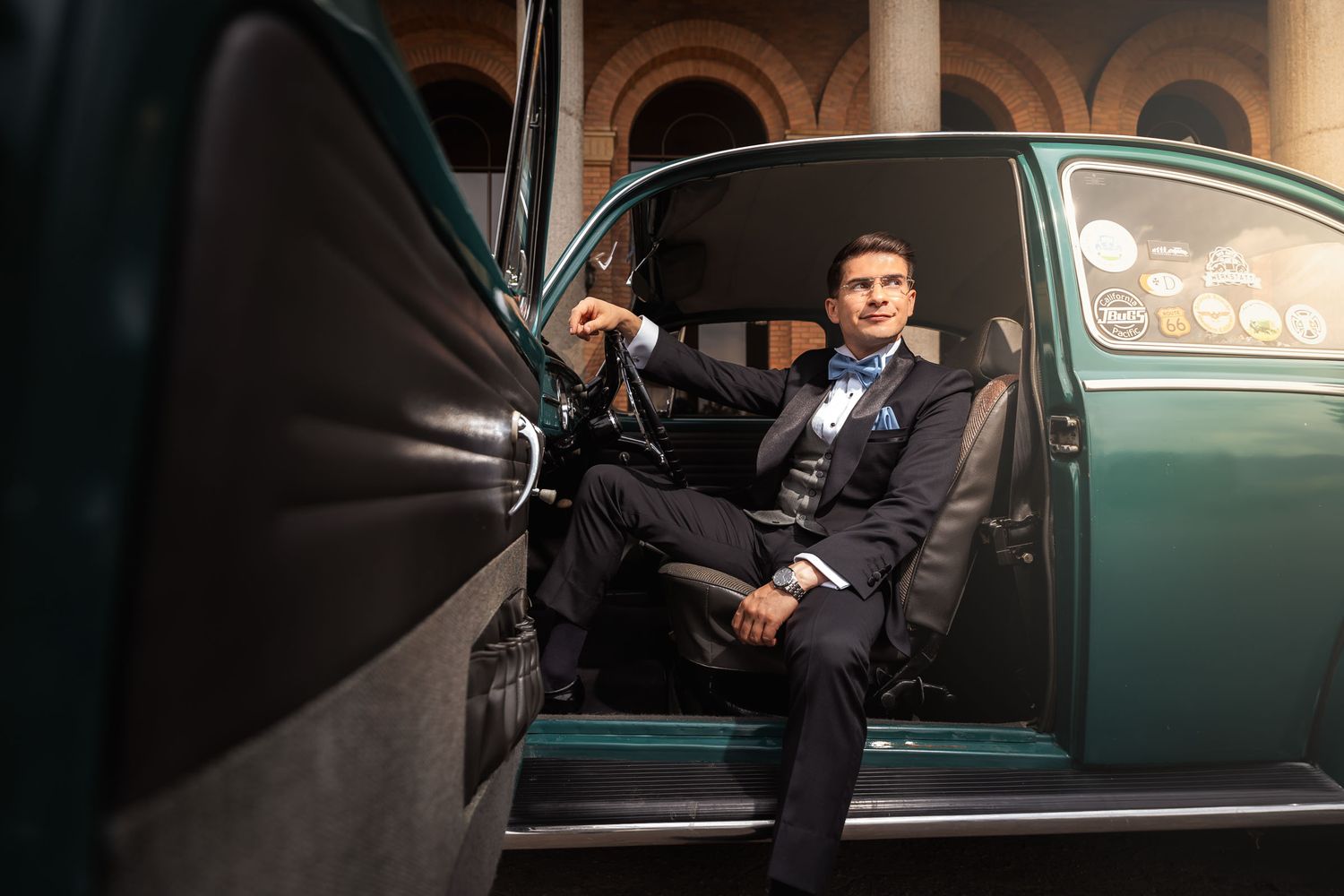 Novio con esmoquin y pajarita azul posa sentado al volante de coche VW Escarabajo verde; puerta abierta. Fotografía boda.