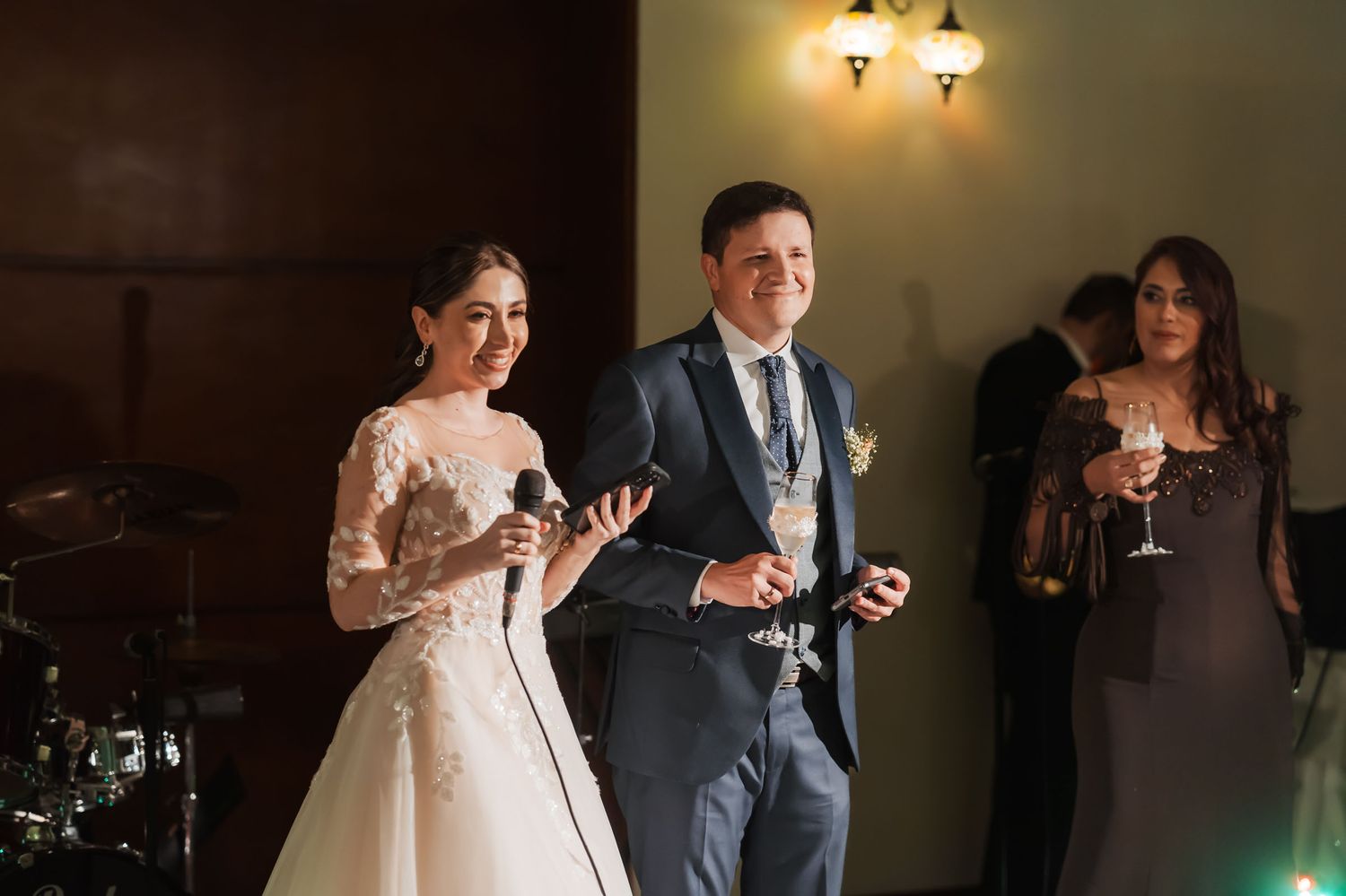 Novia con micrófono y novio con copa sonríen dando discurso o brindis durante recepción de boda en salón.