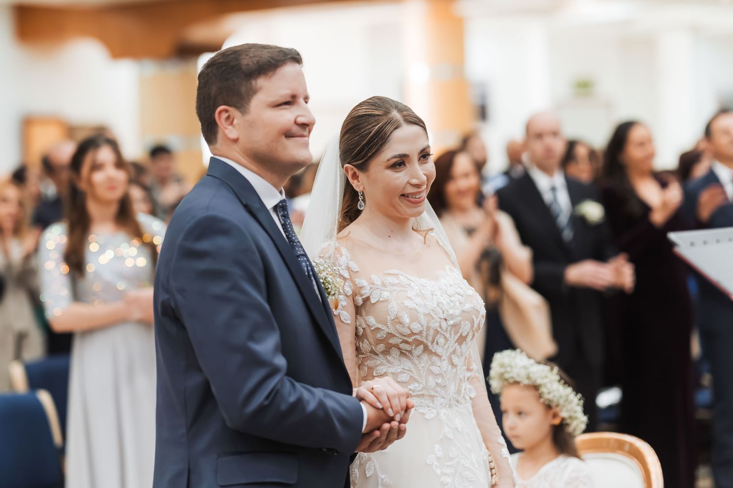 Novios sonrientes tomados de la mano durante ceremonia de boda; invitados aplaudiendo al fondo. Boda IDMJI.