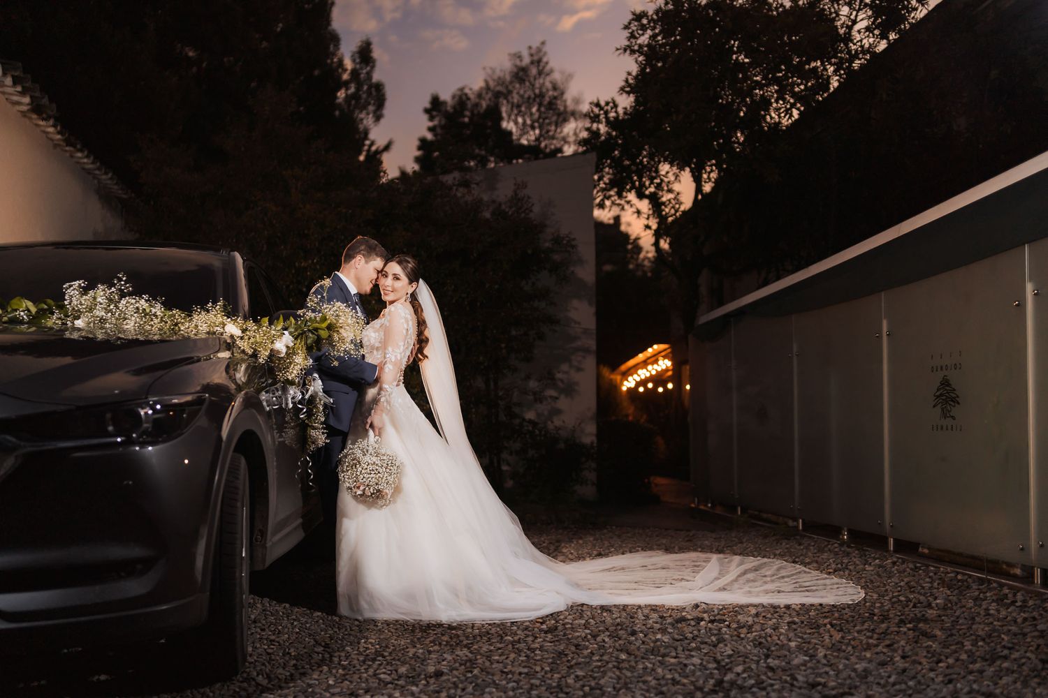 Novios posan abrazados junto a coche oscuro decorado con flores; velo largo de novia se extiende. Foto boda al atardecer.