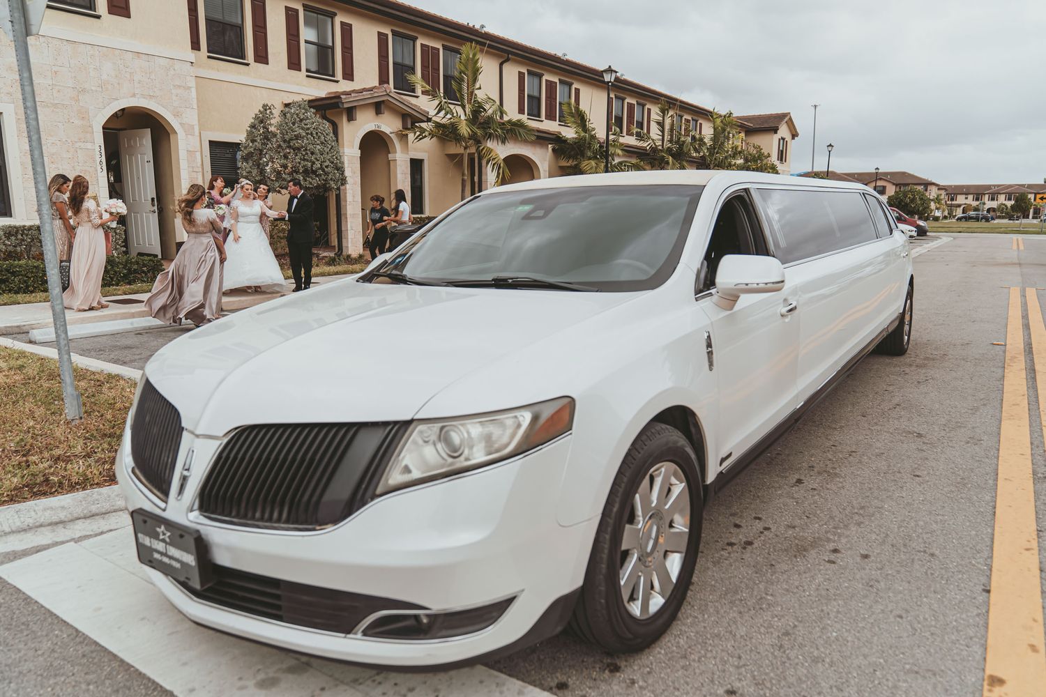 Limusina SUV blanca estacionada en calle; cortejo nupcial (novios, damas) camina al fondo. Transporte boda Miami.