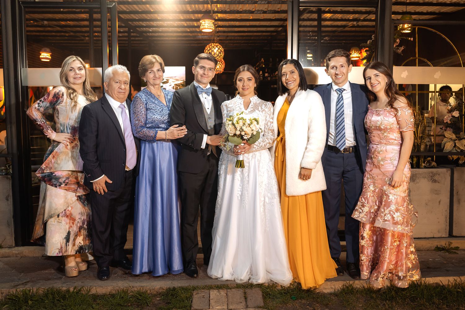 Novios posan felices con su familia en foto de grupo exterior de noche, frente a edificio con luces. Boda.