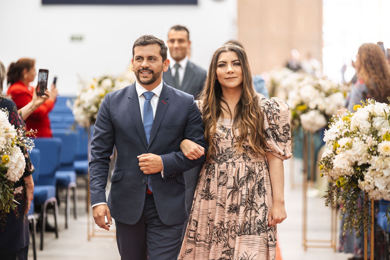 Pareja camina por el pasillo de la iglesia en boda IDMJI; él con traje azul, ella con vestido estampado rosa y negro.