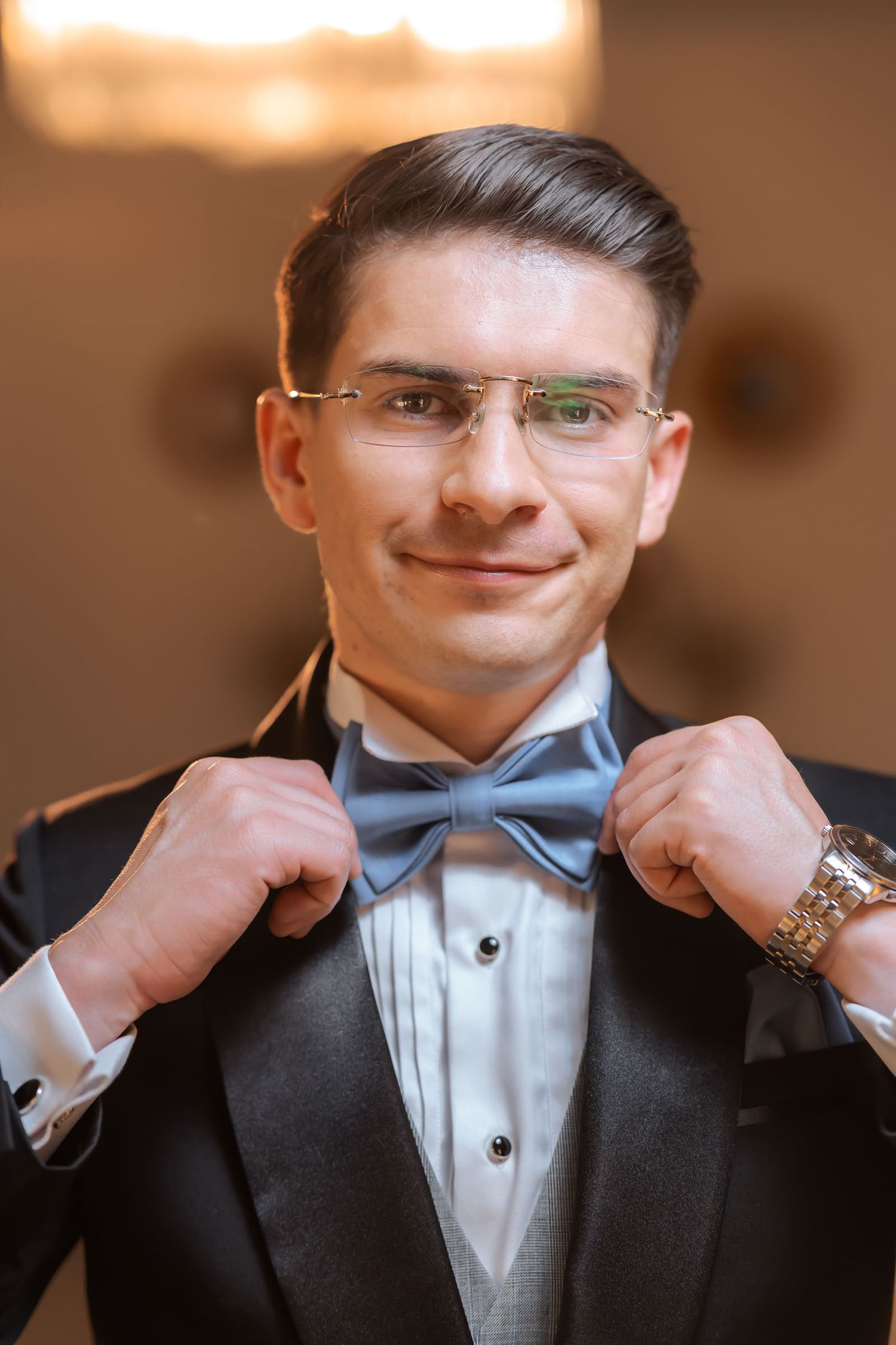 Primer plano: novio sonriente ajustando su pajarita azul; viste esmoquin con chaleco gris. Preparación boda.