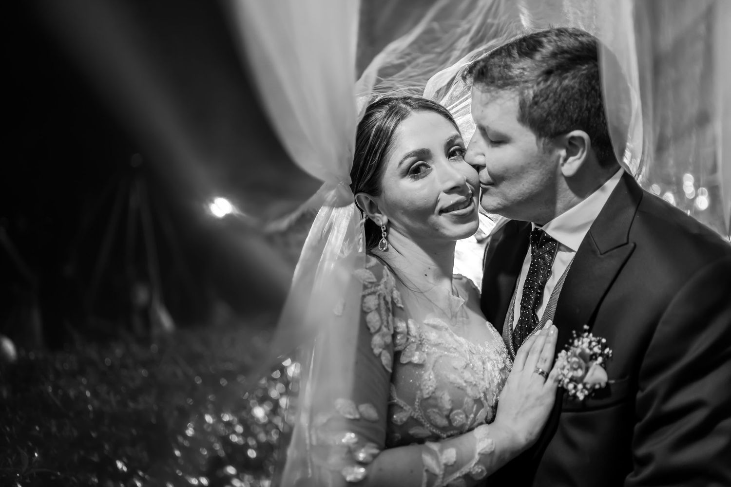 B&W primer plano: Novio besa o susurra a novia sonriente; momento íntimo de boda con fondo suave mientras el velo los cubre.