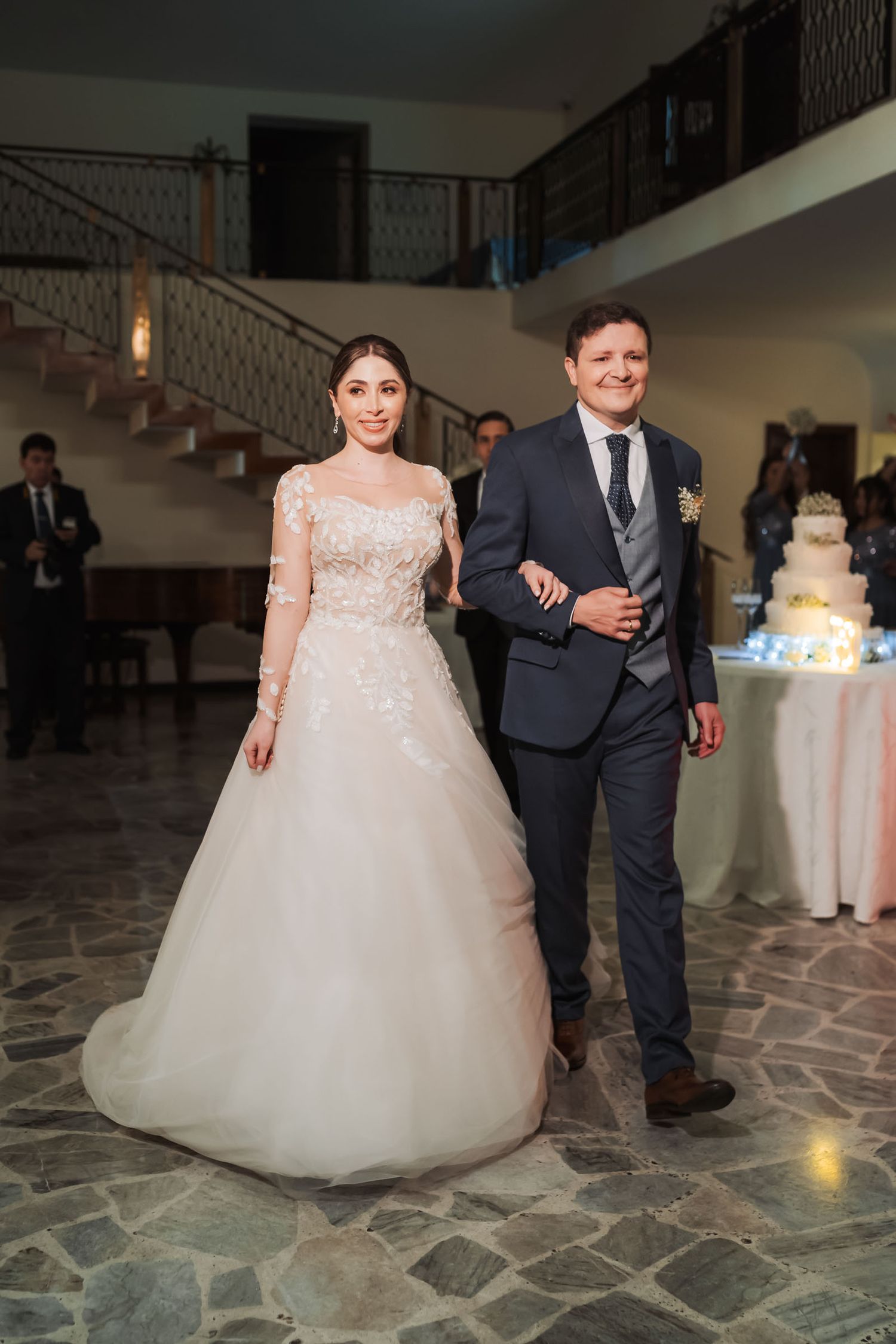 Novia del brazo del novio caminan sonrientes durante recepción de boda; salón elegante con piso de piedra.