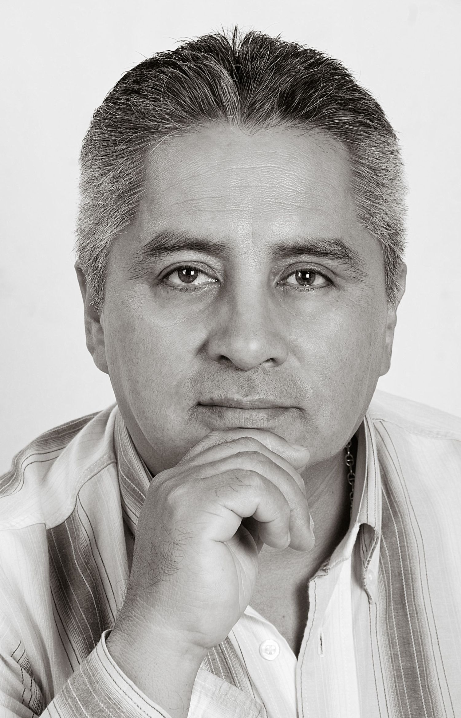 Retrato blanco y negro hombre mediana edad mira serio a cámara, mano en barbilla; camisa rayas. Pensativo.