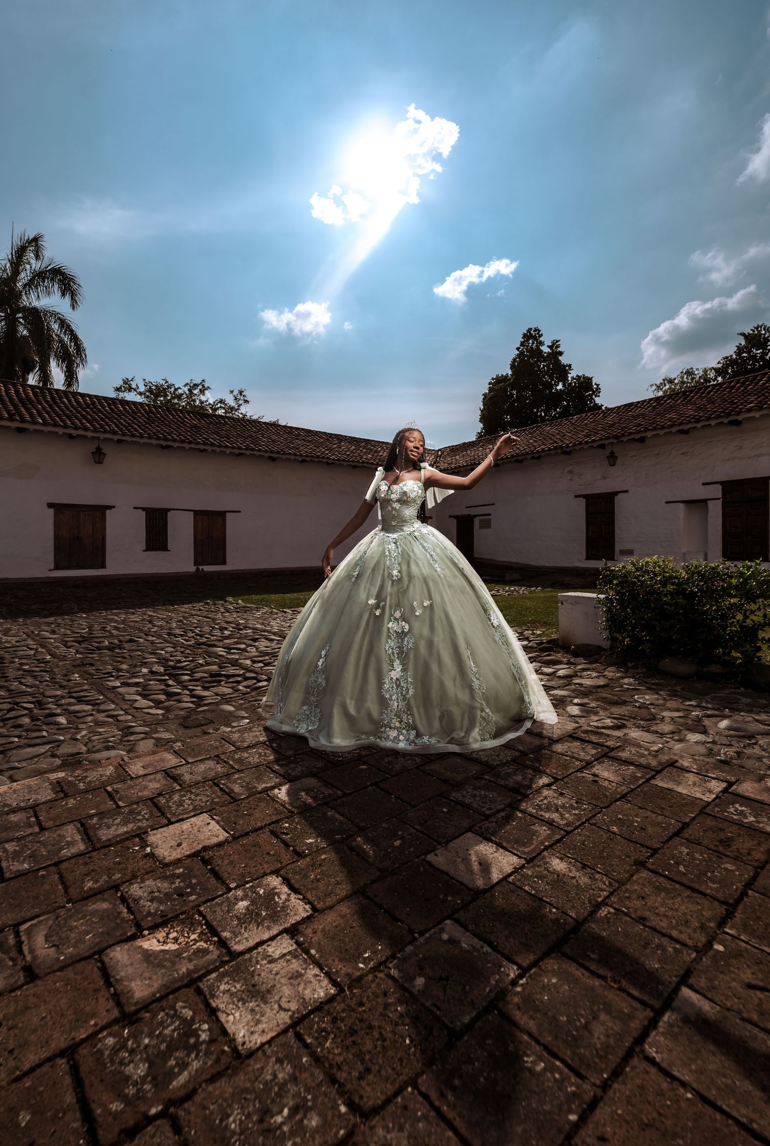 Quinceañera con vestido princesa verde/plata posa en patio adoquinado bajo sol brillante; edificio colonial.