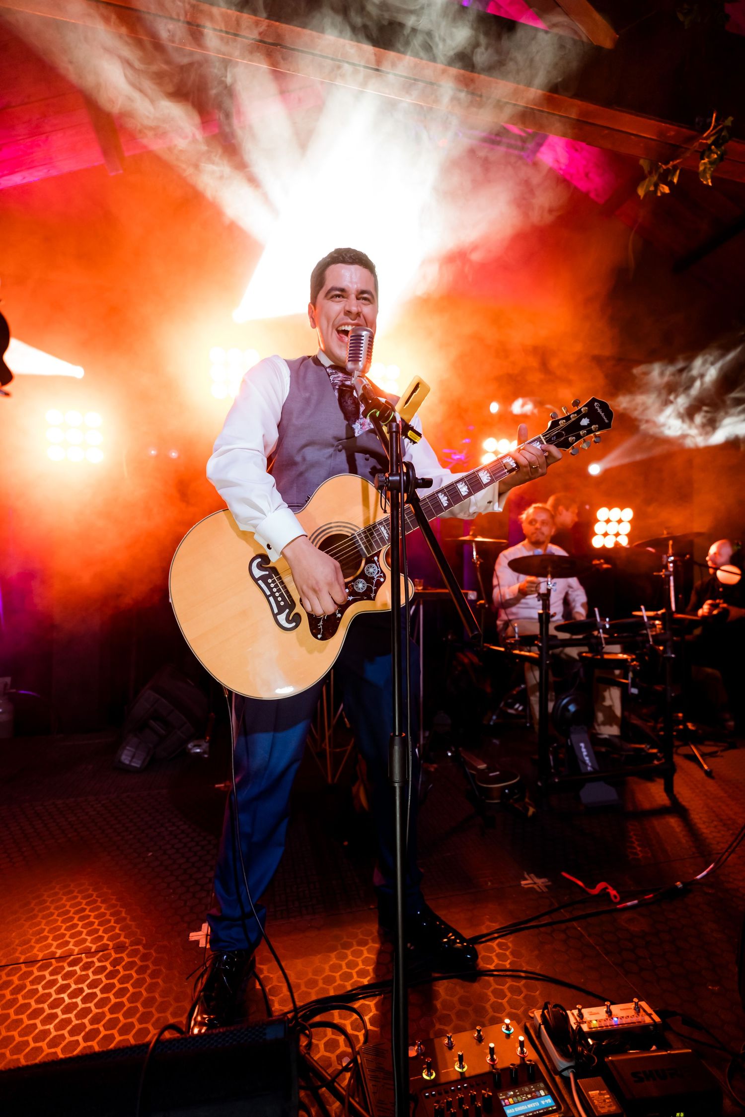 Novio tocando guitarra acústica y cantando en vivo en un escenario iluminado con luces rojas y humo. Viste camisa blanca y chaleco gris