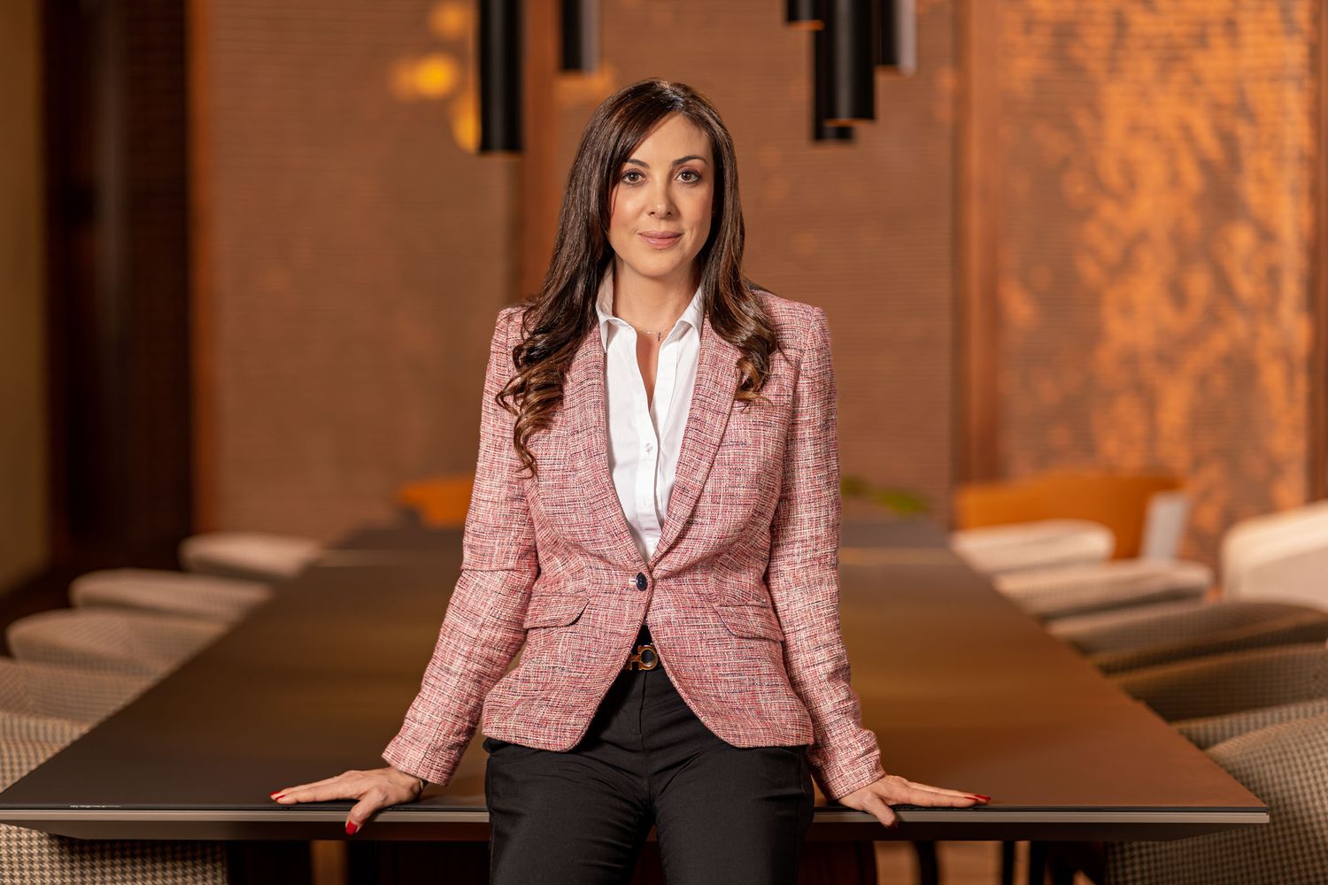 Mujer profesional posa apoyada en mesa de conferencias; lleva blazer tweed rosa, camisa blanca. Entorno oficina. Sandra Vélez