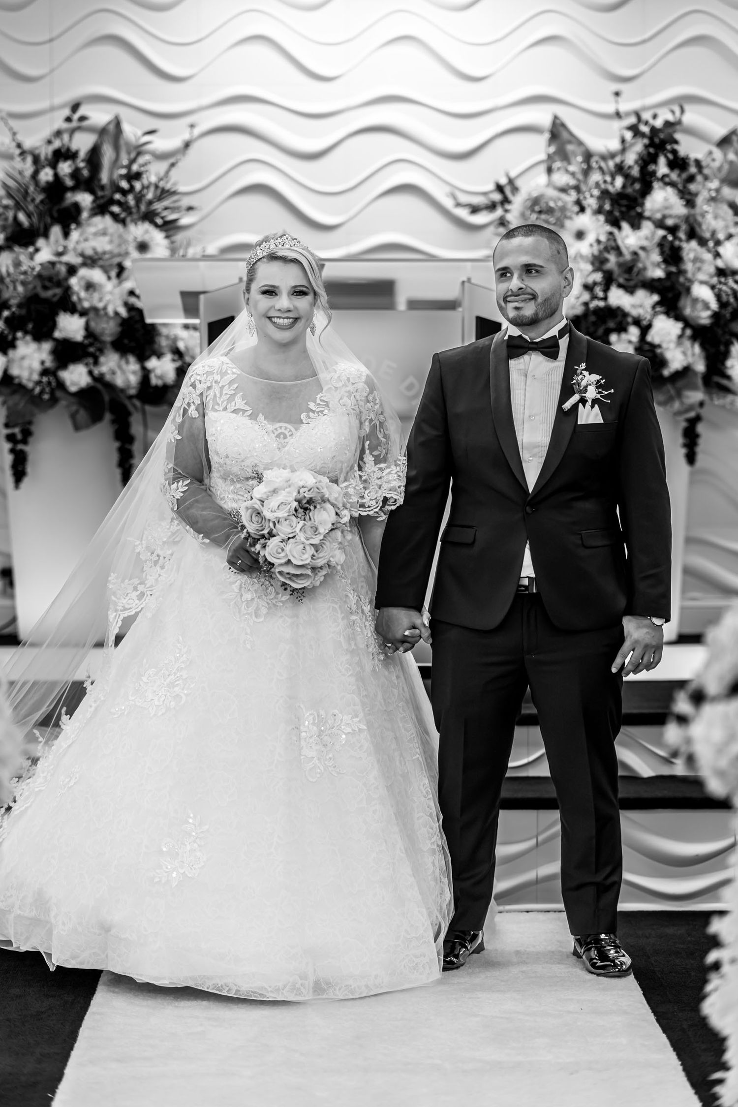Foto B&W: Novios posan tomados de la mano frente al altar de iglesia decorado con flores. Retrato boda.