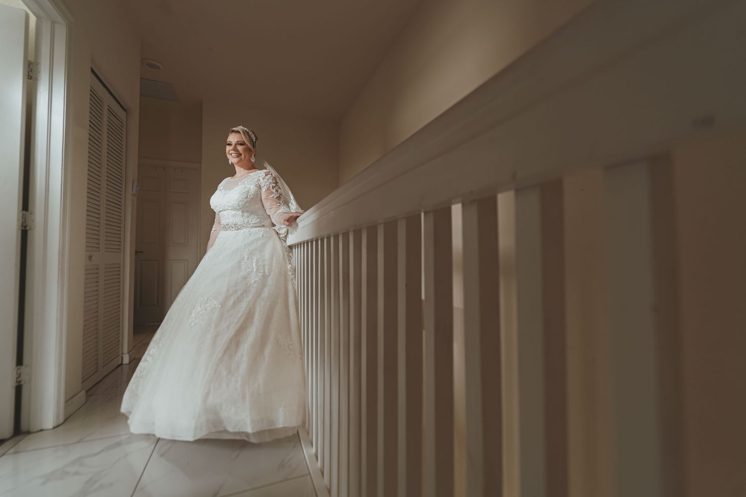 Novia con vestido blanco y velo posa sonriente en puerta de casa junto a su madre (vestido brillante). Preparación boda.