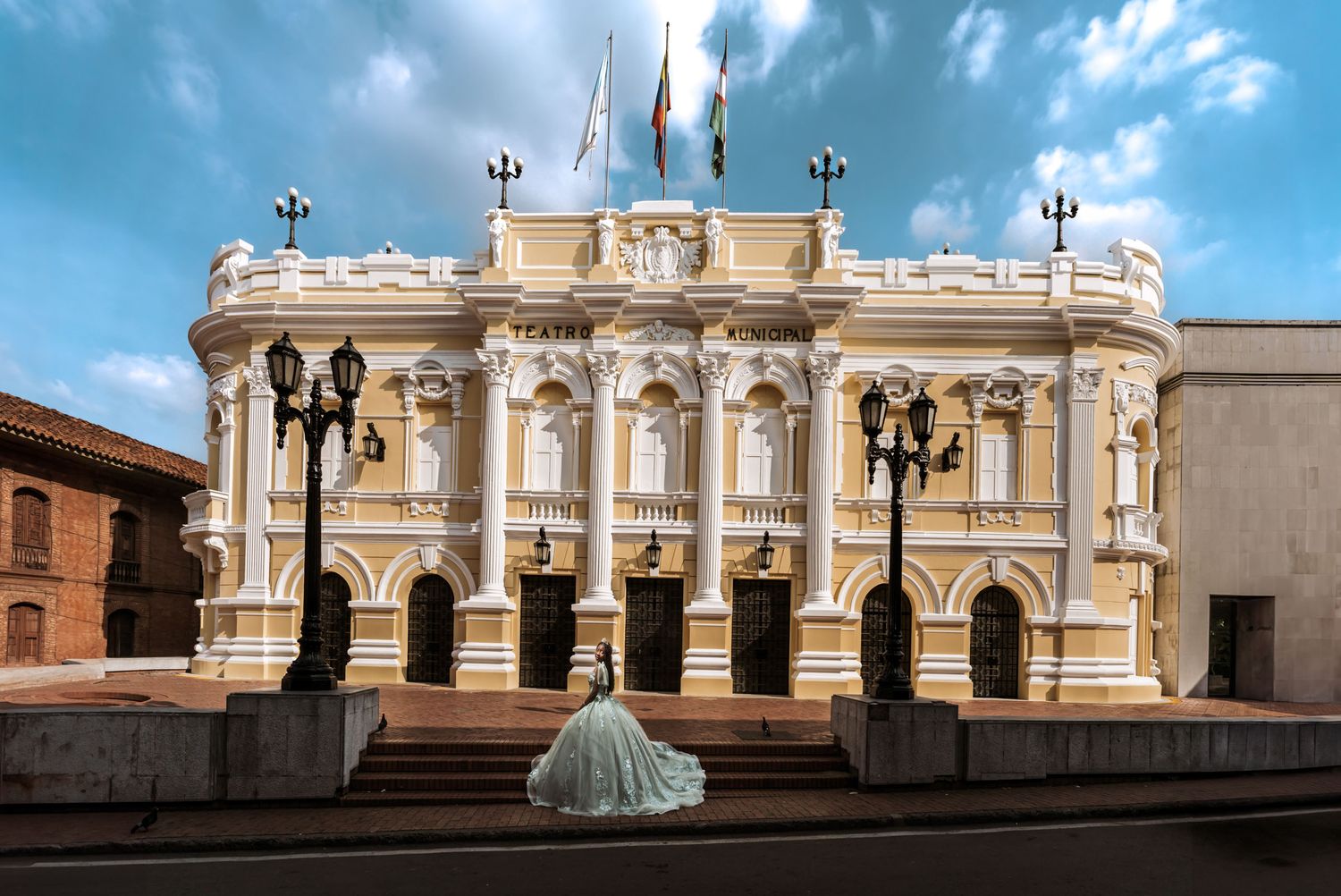 Quinceañera vista de espaldas con vestido princesa verde/plata frente a edificio clásico (teatro?); sesión en exterior.