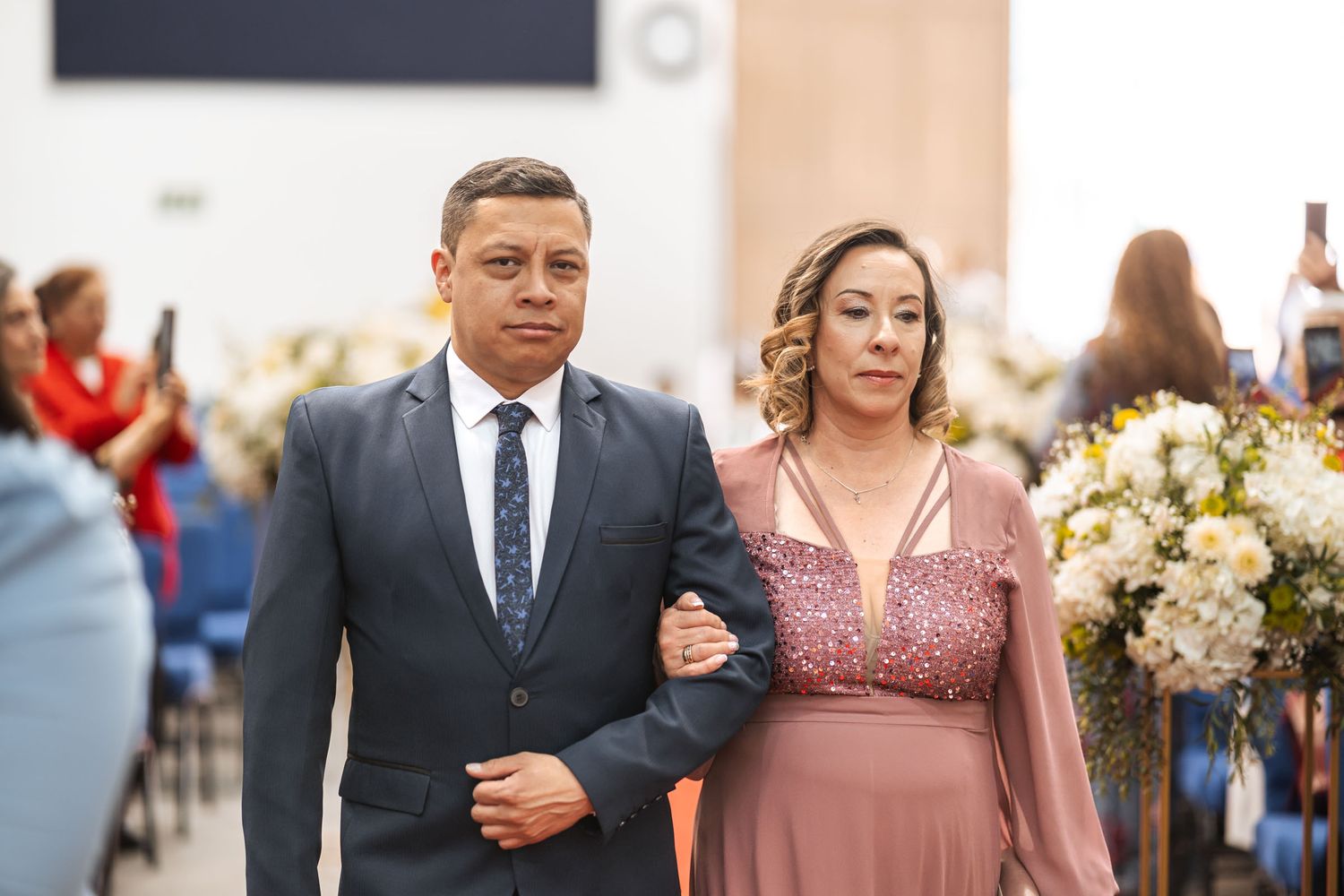 Pareja camina por pasillo de iglesia en procesión de boda; ella con vestido rosa con detalles brillantes, él con traje azul.