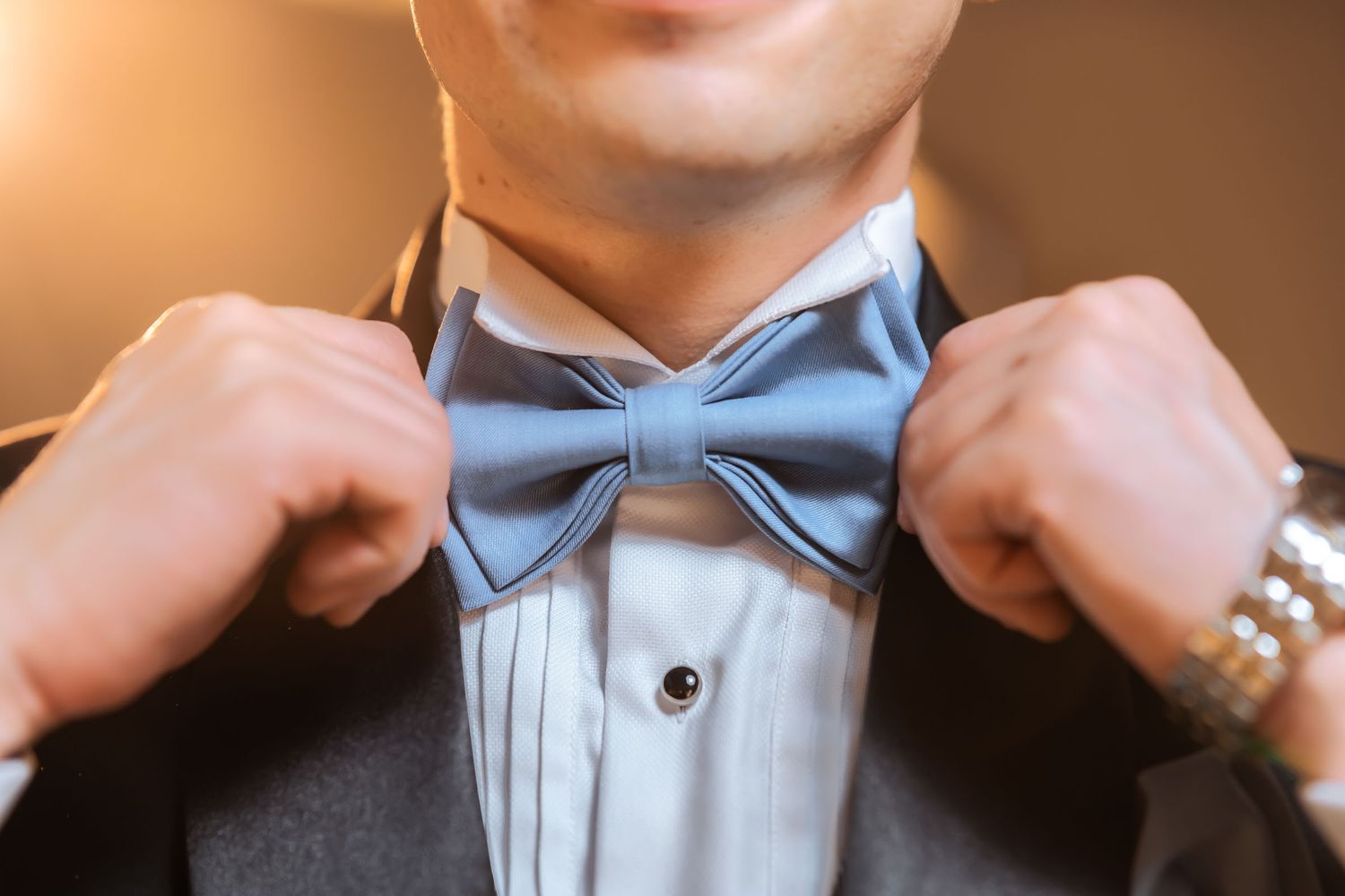 Detalle cercano: Novio ajustando pajarita azul cielo sobre camisa blanca plisada de esmoquin; preparación boda.