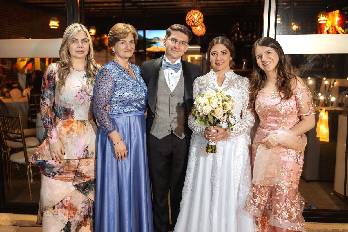 Novios sonrientes posan con familia en recepción de boda exterior nocturna. Grupo elegantemente vestido.