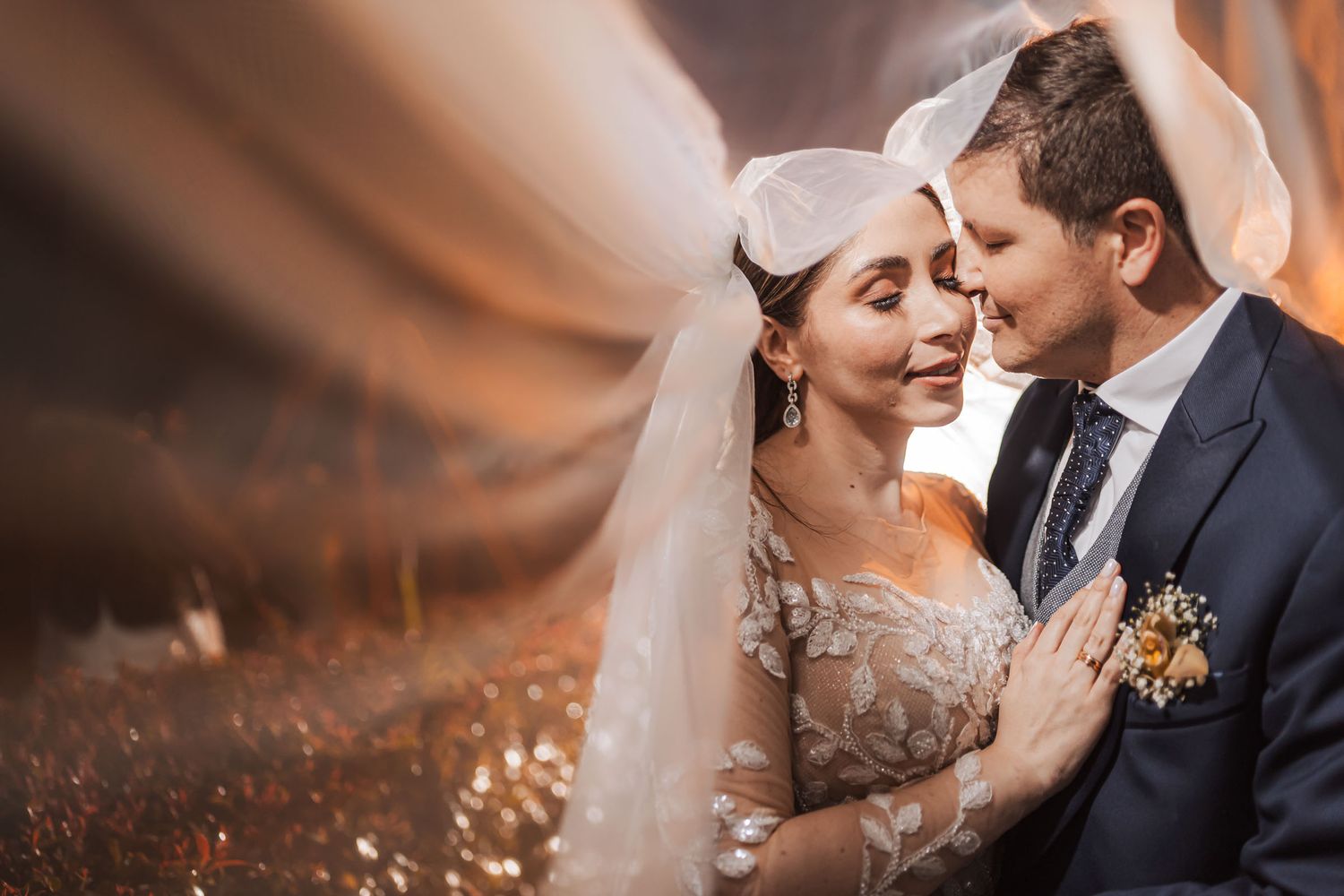 Primer plano íntimo de novios bajo velo transparente; luz cálida, vestido bordado. Momento tierno de boda.