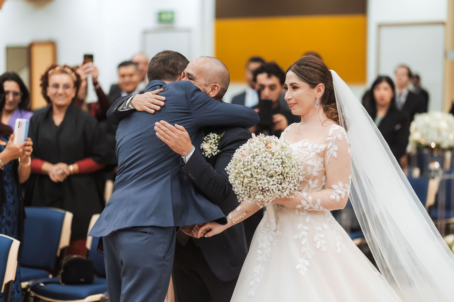 Novia con ramo mira sonriente mientras novio abraza a invitado; momento emotivo post-ceremonia boda.