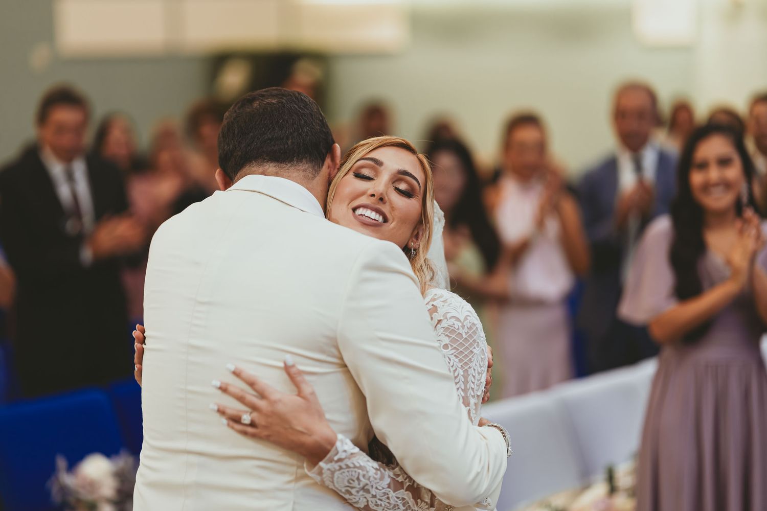 Novia sonriente abraza con ternura al novio (chaqueta blanca) durante recepción; invitados aplauden.