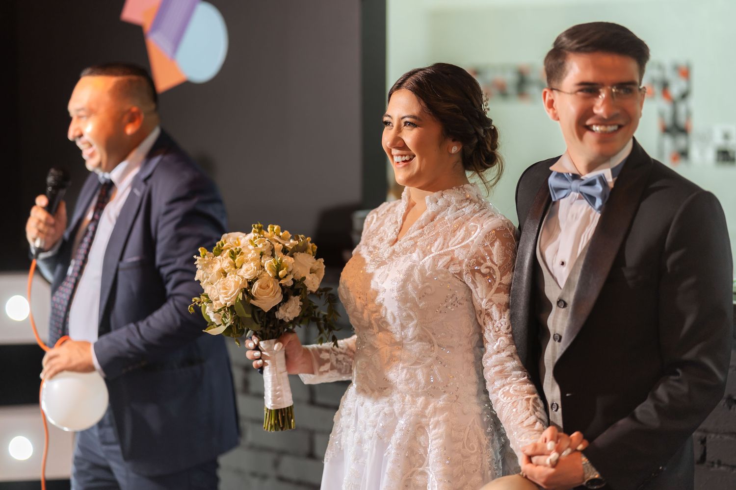 Novios ríen escuchando discurso con micrófono en recepción de boda. Invitado habla, pareja feliz.