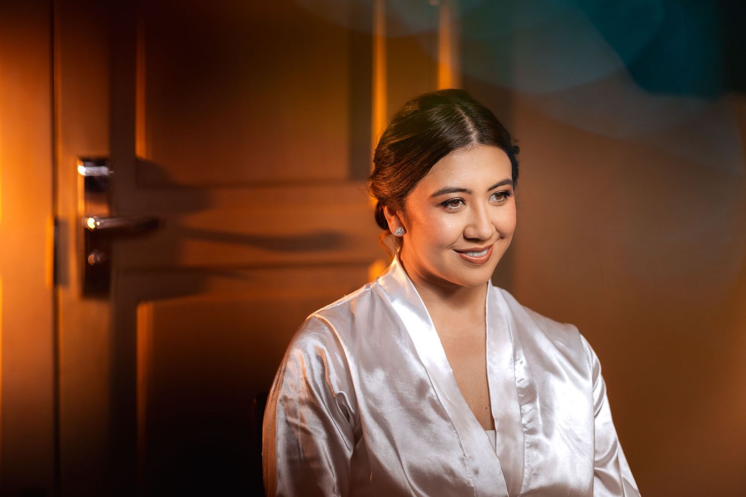 Novia sonriente con bata blanca de satín cerca de una puerta; luz cálida en preparación de boda.