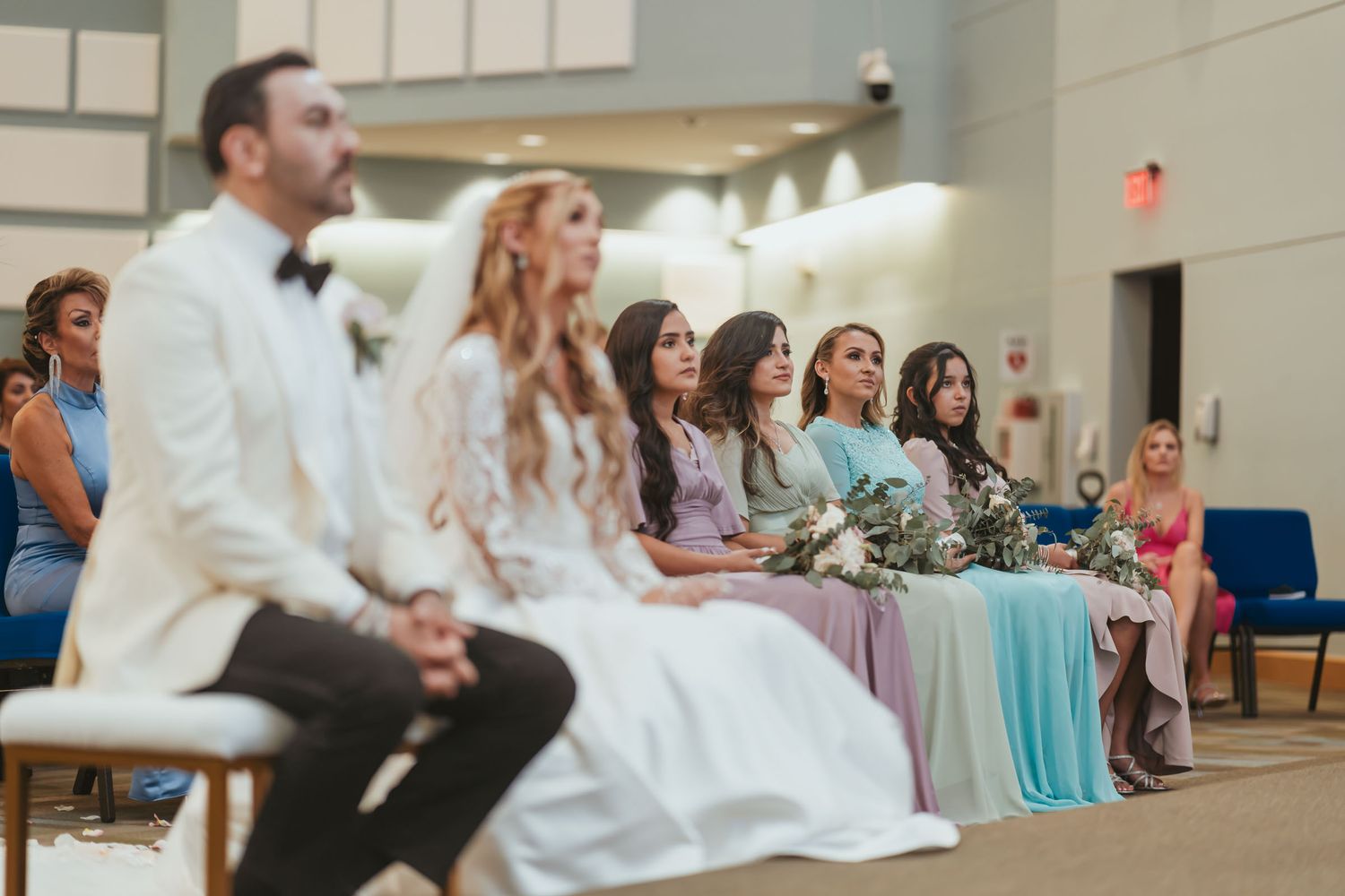 Novios y damas de honor sentados en primera fila durante ceremonia de boda en iglesia/salón.