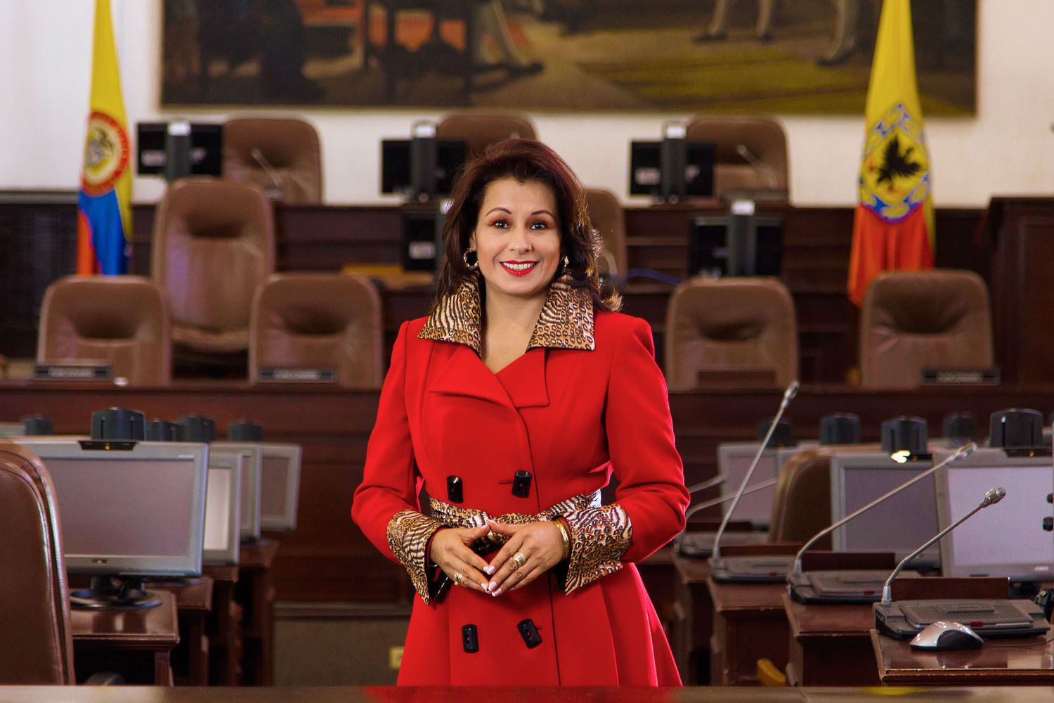 Mujer sonriente con abrigo rojo elegante posa de pie en auditorio o cámara legislativa; bandera Colombia fondo. Olga Victoria Rubio