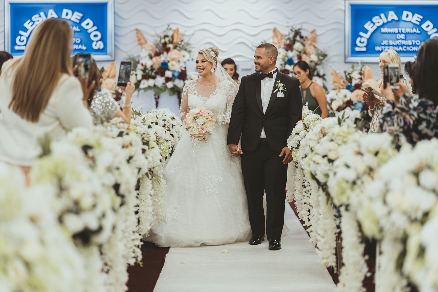 Novios sonrientes desfilan por pasillo de iglesia con flores blancas, invitados les fotografían. Momento de alegría en boda IDMJI