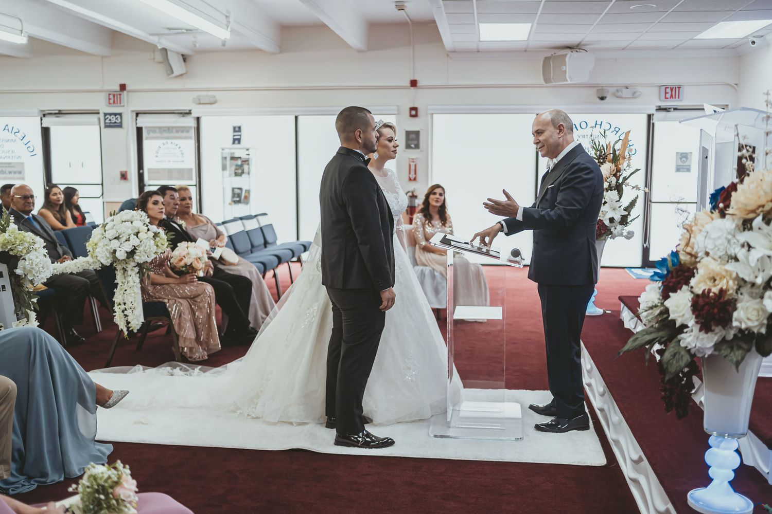 Novios escuchan al oficiante durante ceremonia de boda en altar; invitados sentados al fondo.