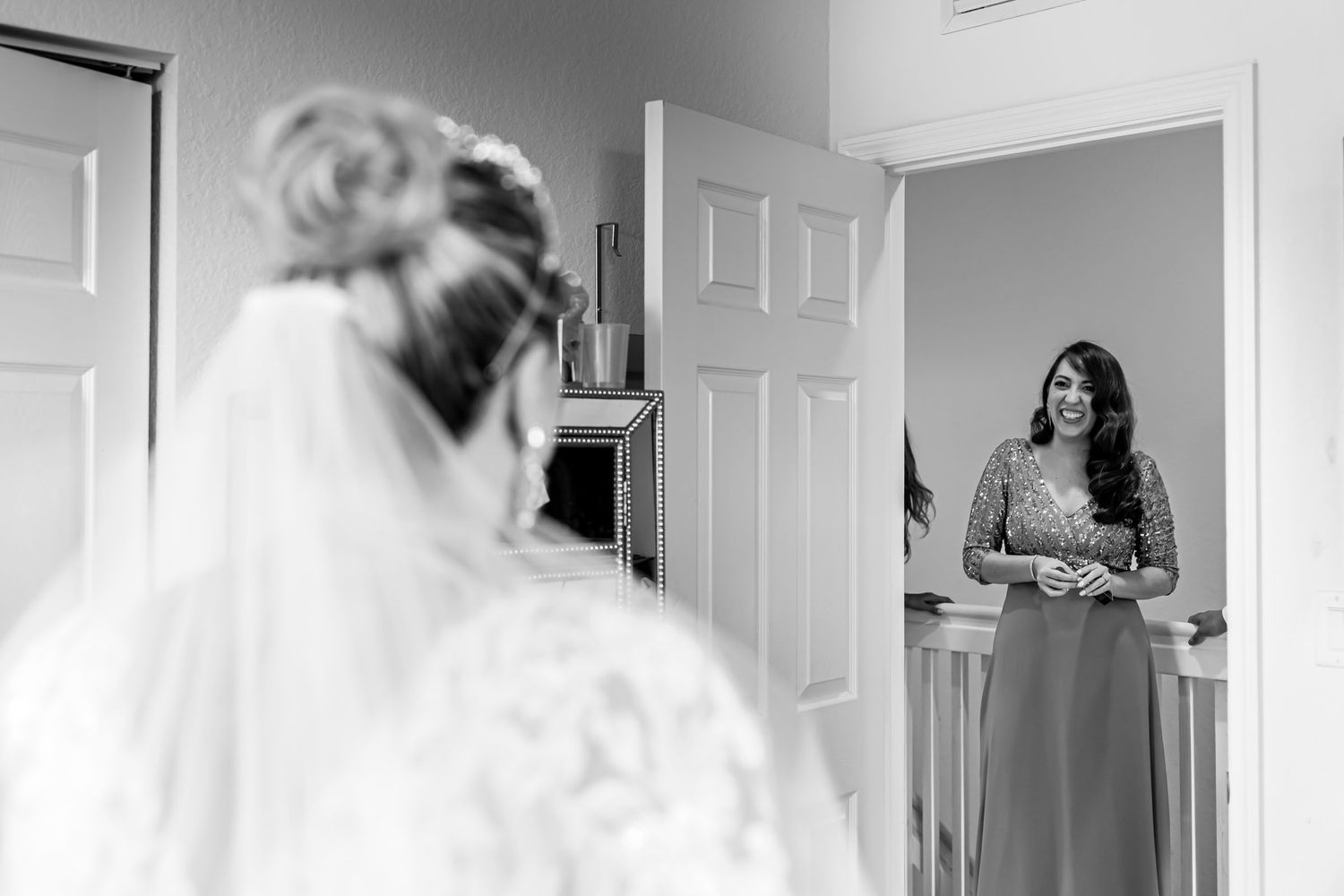Foto B&W: Mujer sonríe conmovida al ver a novia (vista espalda) por primera vez vestida; 'first look' boda.