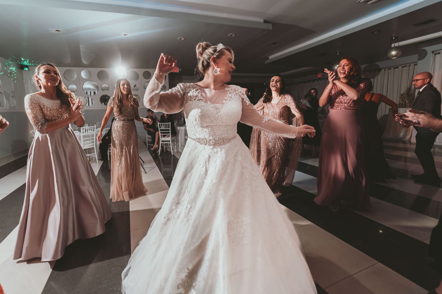 Novia baila feliz en centro de pista a cuadros rodeada de invitados que aplauden; fiesta boda.