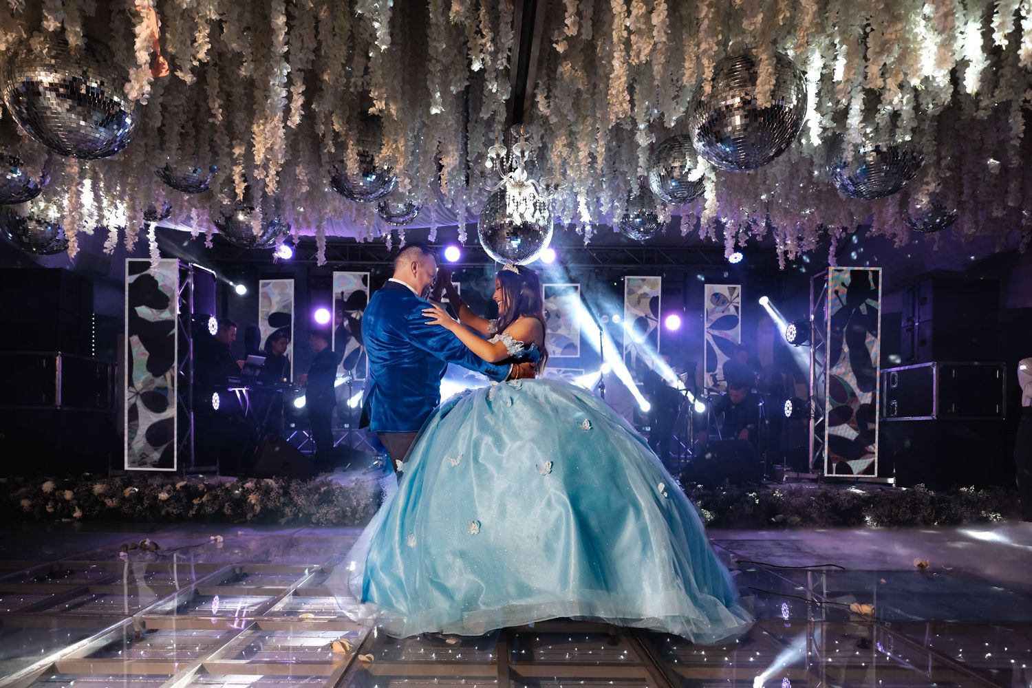 Quinceañera (vestido azul) baila vals con padre/escolta en salón elegante; techo decorado, luces, pista.