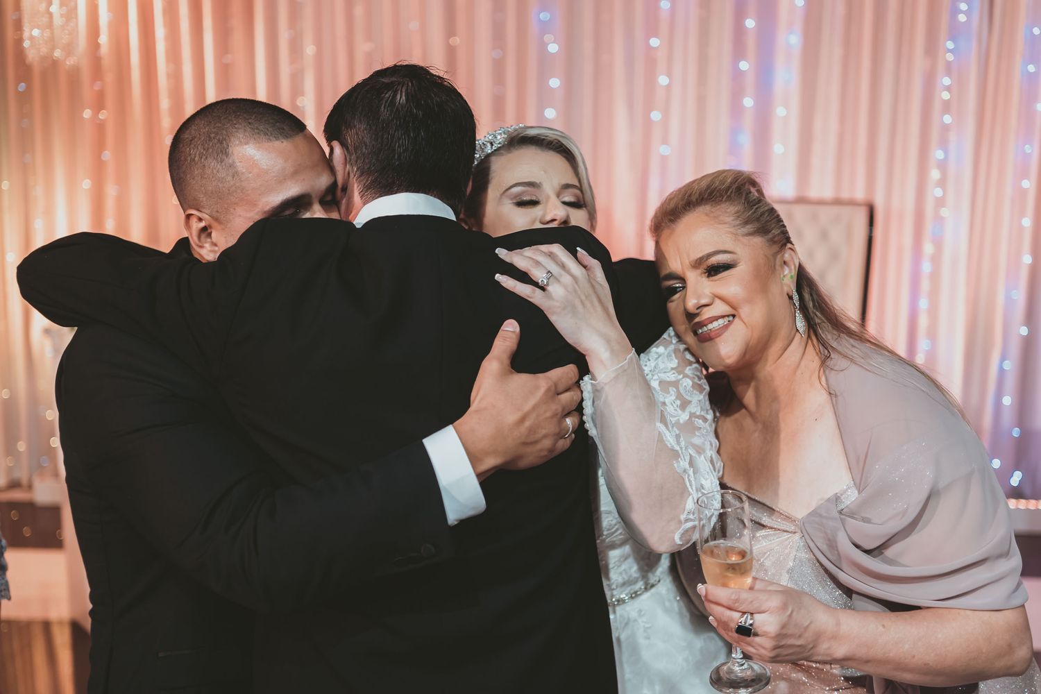 Novios comparten abrazo grupal emotivo con familiares o invitados durante recepción de boda; fondo luces.